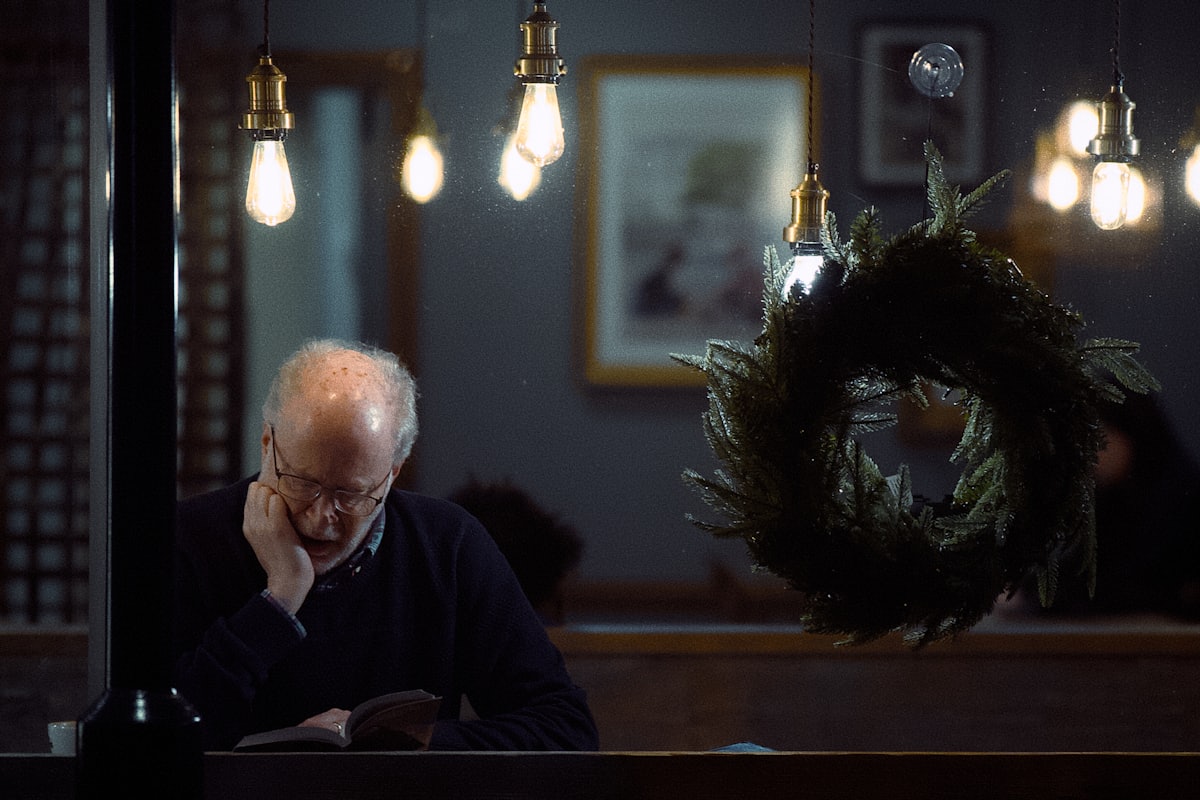 An older man reading alone at a table in a dim room lit by hanging bulbs