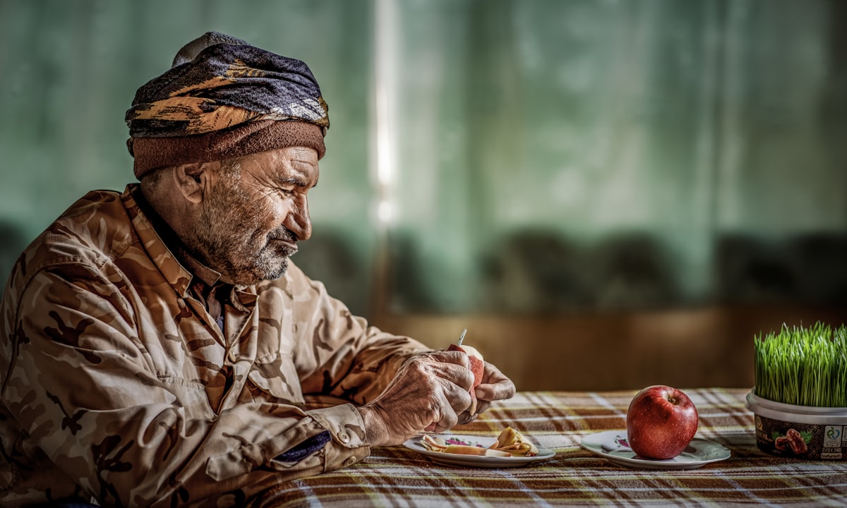 An older man in a head wrap eating a simple meal at a table set with an apple and a small plant
