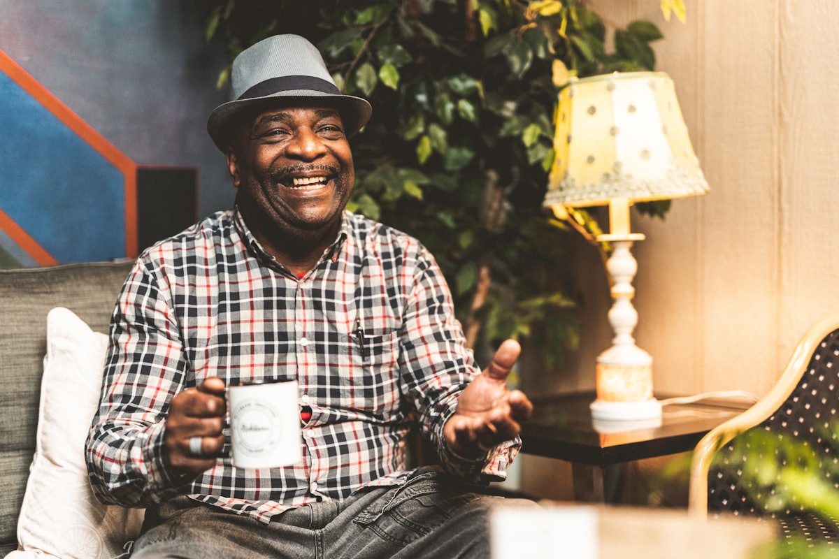 An older man in a grey trilby and plaid shirt laughing on a sofa, a mug of tea in one hand