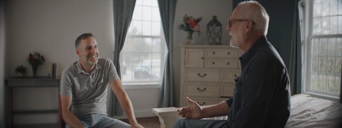 An older man and a younger man sitting talking on the edge of a bed in a bright bedroom