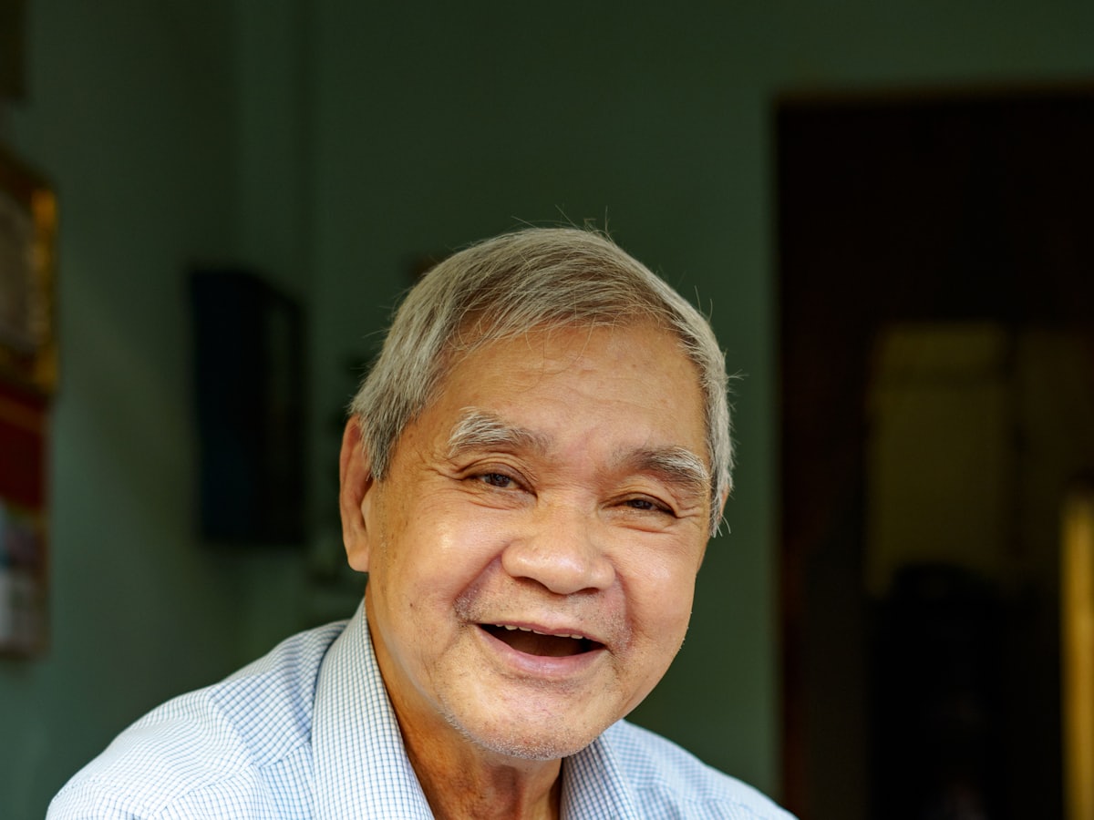 A close portrait of a smiling older man with grey hair in a pale checked shirt, at home