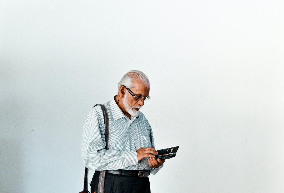 An older man with a white beard and a bag over his shoulder, standing against a blank wall looking at his phone