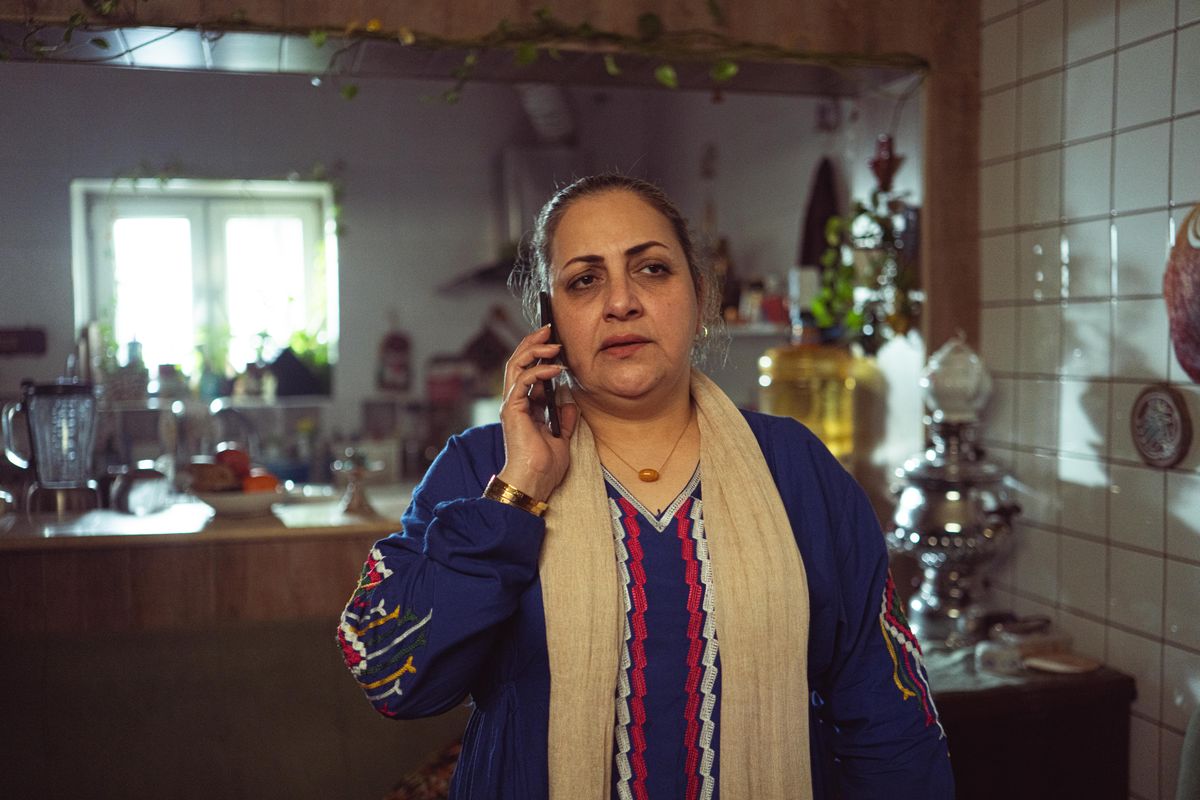 A woman in her fifties standing in her kitchen with a phone held to her ear, listening with a serious expression
