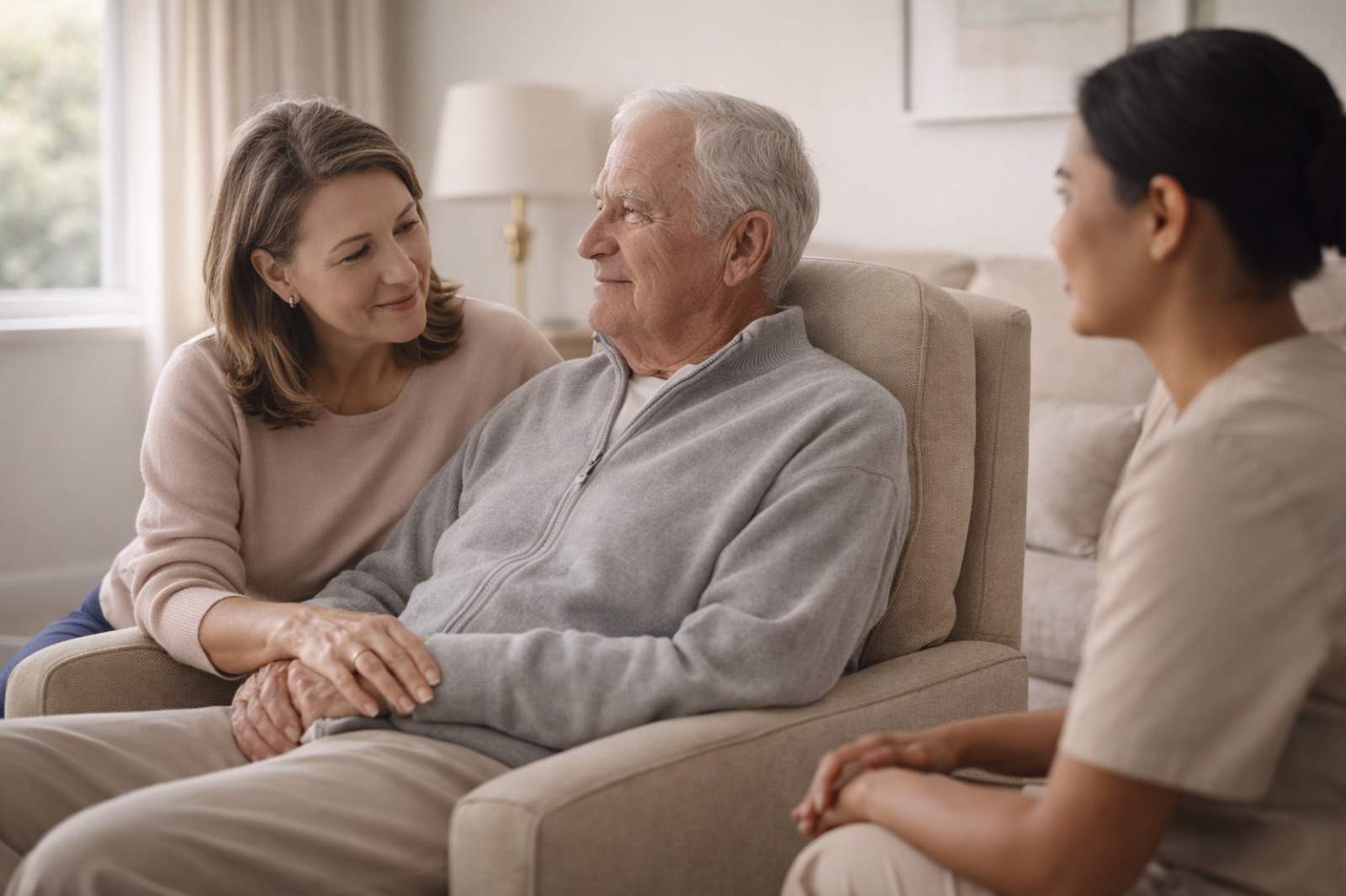 An older man seated in an armchair at home, his adult daughter beside him and a caregiver seated across from them, mid-conversation